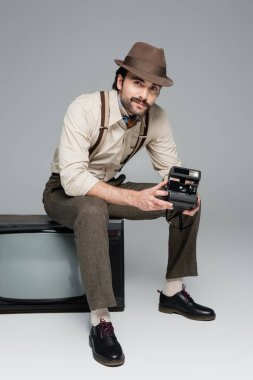 full length of smiling man retro style clothing and hat holding vintage camera and sitting on antique tv on grey  clipart