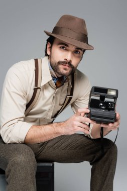 Eski moda kıyafetler ve eski model bir kamera taşıyan şapka ve gri TV 'de izole edilmiş antika bir TV' de oturan adam. 