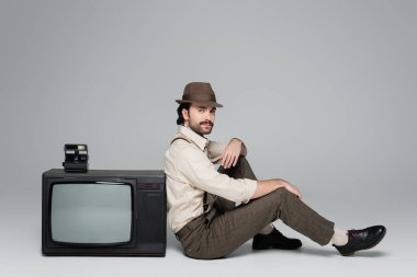 Tam boy erkek retro tarzı kıyafetler ve antika televizyonun yanında oturan gri renkli kameralı bir şapka. 