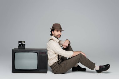 full length of man retro style clothing and hat sitting near antique tv with vintage camera on grey  clipart