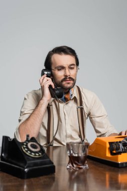Gözlüklü gazeteci, retro telefonda daktilonun yanında konuşuyor ve gri üzerine izole edilmiş bir bardak viski.