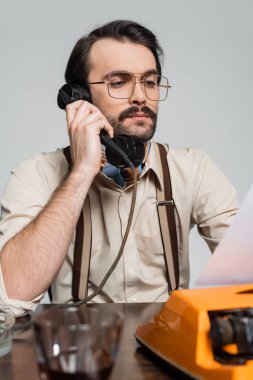 Gözlüklü gazeteci retro telefondan konuşuyor ve gri üzerine izole edilmiş daktiloyla kağıda bakıyor.