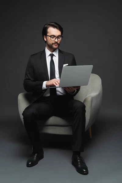 スーツや眼鏡を着たビジネスマンがアームチェアに座って暗い灰色のノートパソコンを使って — ストック写真
