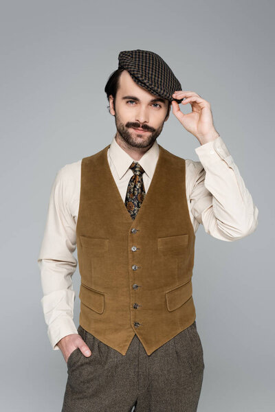 man with mustache and retro clothing posing with hand in pocket while adjusting vintage hat isolated on grey