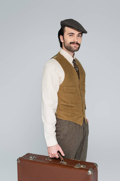 smiling man with mustache and retro style clothing standing with luggage isolated on grey