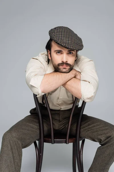 stock image man with mustache in retro style clothing and hat sitting on wooden chair on grey 