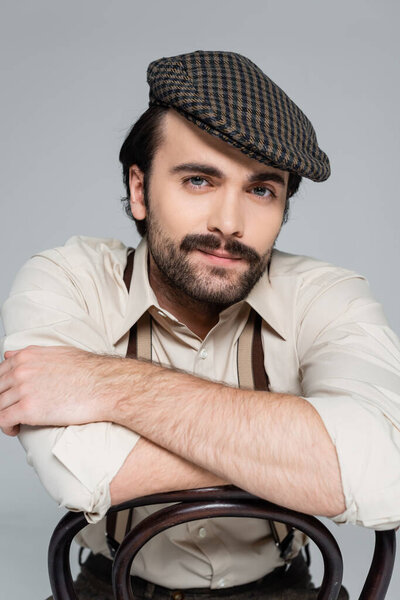 человек с усами в ретро-стиле одежды и шляпы сидя на деревянном стуле на сером 