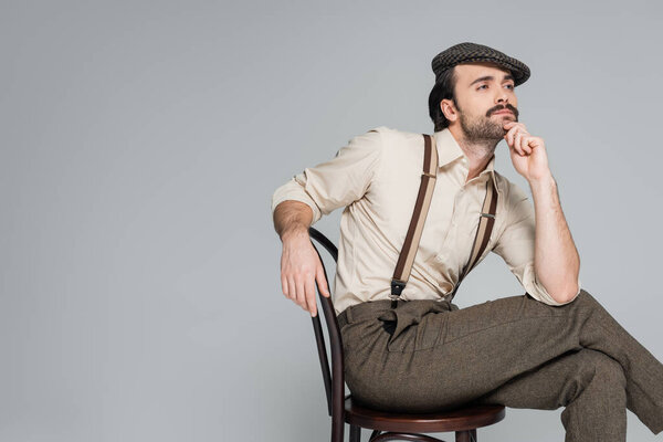 pensive man with mustache in retro style clothing and hat sitting on wooden chair isolated on grey 