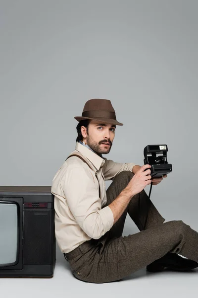 男子复古风格的服装和帽子坐在古董电视机旁边 灰色的老式相机 — 图库照片