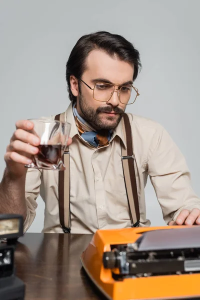 戴眼镜的记者一边拿着玻璃杯 一边拿着灰色的威士忌 一边用打字机看报纸 — 图库照片