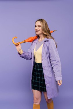 positive young woman in trench coat standing with orange umbrella isolated on purple  clipart