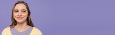 sly young woman looking away and smiling isolated on purple, banner clipart