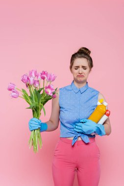 Plastik eldivenli hoşnutsuz genç bir kadın elinde deterjanlı şişeler ve pembe üzerine izole edilmiş laleler tutuyor.