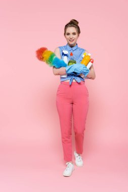 Tam boy lastik eldivenli, elinde deterjanı olan ve pembe üzerine toz fırçası süren genç bir kadın.