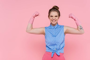 happy and tattooed woman in rubber gloves showing power isolated on pink  clipart