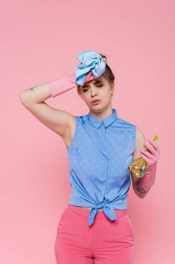 Lastik eldivenli yorgun genç bir kadın. Elinde mavi bir bez parçası ve pembe bir sprey var. 