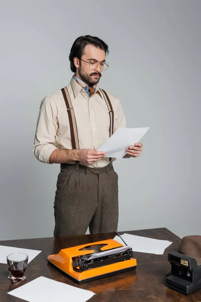 journalist in eyeglasses looking at paper near typewriter, vintage camera and glass of whiskey isolated on grey