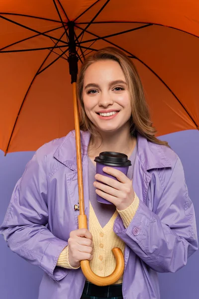 トレンチコートの幸せな若い女性は紙コップを保持し 紫色に隔離されたオレンジ色の傘の下に立って — ストック写真