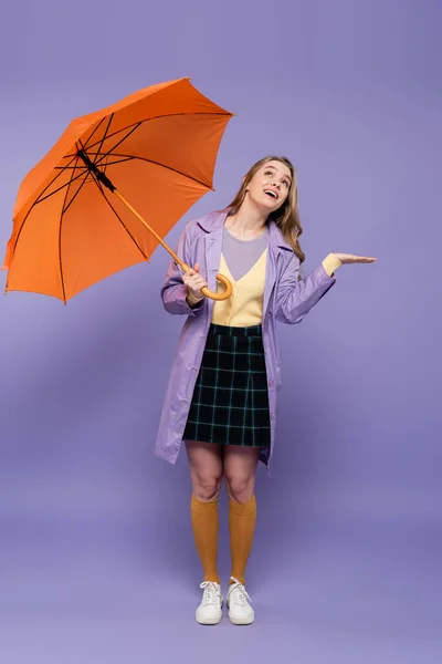 全身上下都是开朗的年轻女子 身穿战壕外套 头戴紫色的橙色雨伞 摆着姿势 — 图库照片