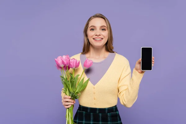 ピンクチューリップを持っている陽気な若い女性と紫に隔離された空白の画面でスマートフォンを示しています — ストック写真