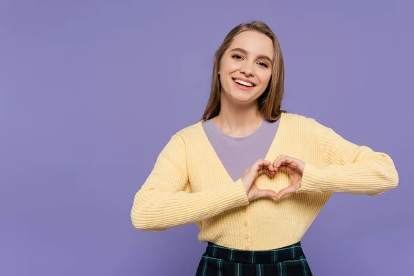 joyful young woman showing heart sign with hands isolated on purple