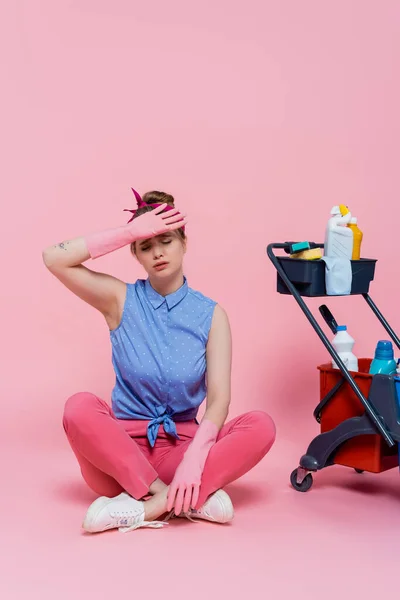 ゴム手袋の疲れ若い女性の完全な長さピンクの上に掃除用品とハウスキーピングカートの近くに座って — ストック写真