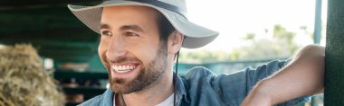 portrait of young bearded farmer in brim hat smiling on farm, banner clipart