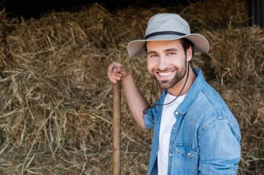 joyful farmer in brim hat looking at camera near blurred haystack on farm clipart