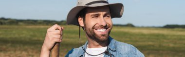 young farmer in brim hat smiling and looking away in field, banner clipart
