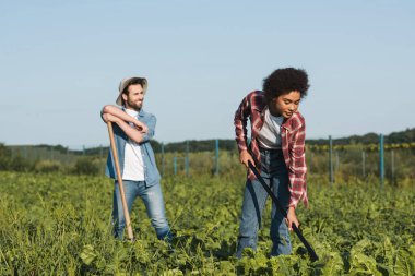 smiling farmer looking away while african american woman working in field clipart