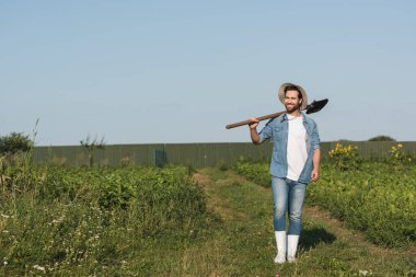 full length view of happy farmer walking with shovel on green field  clipart