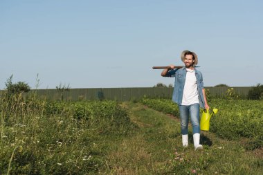 full length view of positive farmer with shovel and watering can walking in field clipart