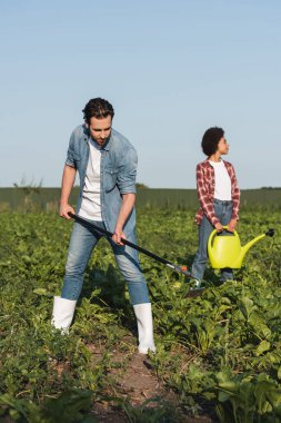 full length view of farmer working with hoe near african american woman with watering can clipart