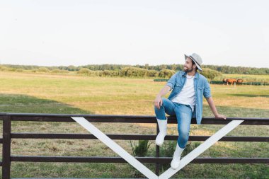 full length view of farmer in denim clothes and brim hat sitting on wooden fence on farmland clipart