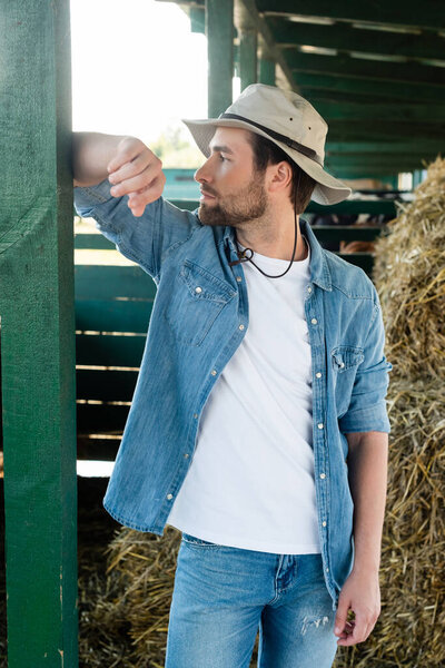 farm worker in denim shirt and brim hat looking away while standing near barn