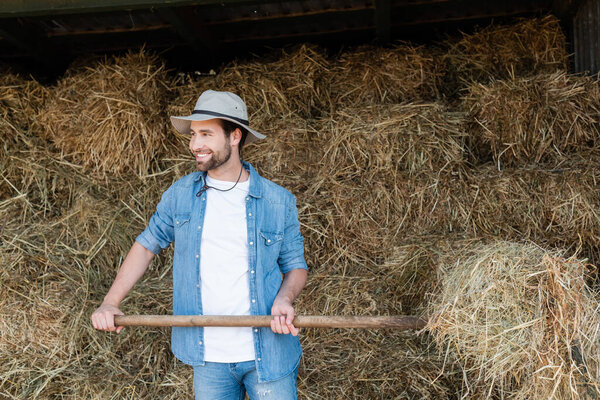 smiling farmer in denim clothes and brim hat looking away while working near haystack