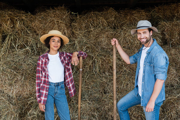 young interracial farmers in hats smiling at camera near haystack