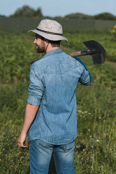 back view of farmer in denim shirt and brim hat standing with shovel in field