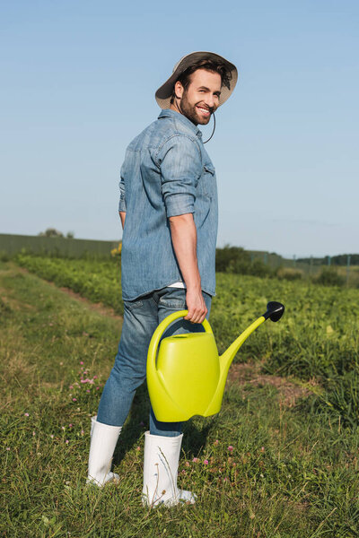 full length view of happy young farmer standing with watering can in field