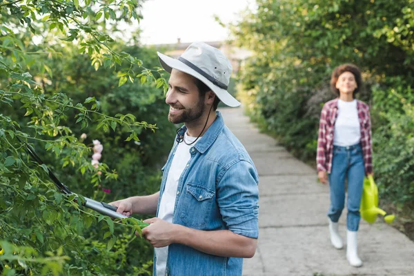 happy gardener trimming bushes near blurred african american woman with watering can