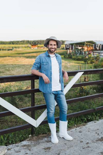 full length view of smiling farmer in denim clothes standing near wooden fence on farm