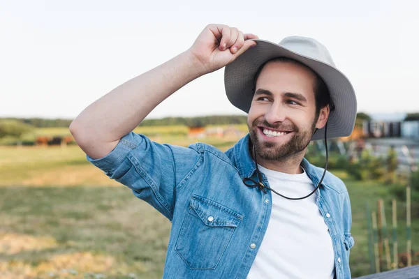 Young Happy Farmer Touching Brim Hat While Looking Away Field — Stock Photo, Image