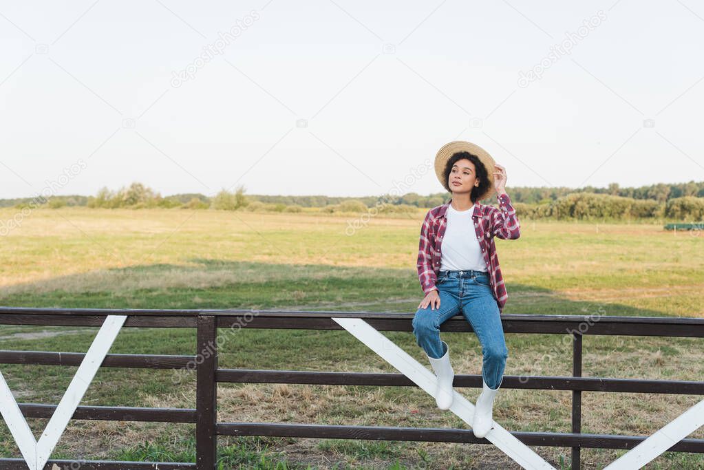 full length view of african american woman looking away while sitting on wooden fence in field