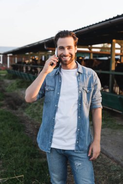 Çiftçi akıllı telefondan konuşuyor. Bulanık kovboy kulübesi yakınlarında ve kameraya gülümsüyor.