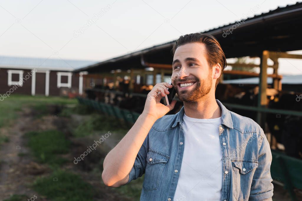 happy farmer looking away while talking on smartphone near blurred cowshed