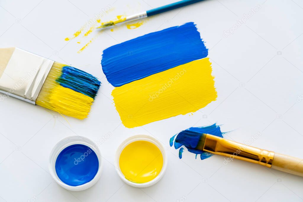 Top view of painted ukrainian flag near paints and paintbrushes on white background 
