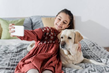 Neşeli kız yatak odasında Labrador 'la cep telefonundan selfie çekiyor.