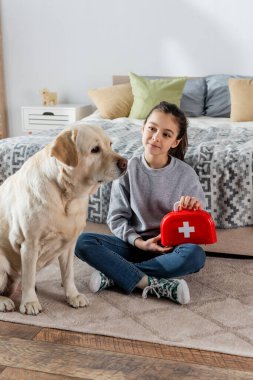 Labrador yakınlarında yerde otururken elinde oyuncak ilk yardım çantası tutan pozitif bir kız.