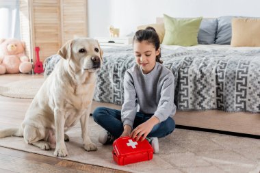 Oyuncak ilk yardım çantasıyla doktorculuk oynayan gülümseyen kız ve yatak odasında yerde labrador köpeği.