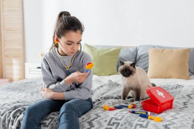 Kedi yakınındaki yatakta oynarken oyuncak steteskop tutan kız.